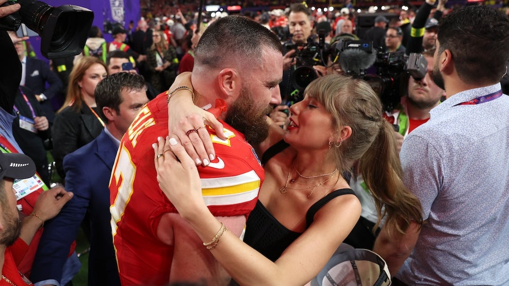 A man on a football field embraces a woman on the field. He wears a red jersey while she wears a black top. They are surrounded by members of the media and others in front of a large crowd.