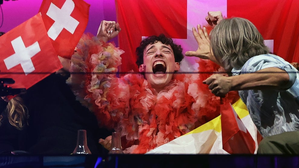 A man wearing a top covered in feathers joyfully celebrates in front of a Swiss flag, surrounded by supporters.