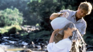 A standing man pours water through a sitting woman's hair, outdoors.