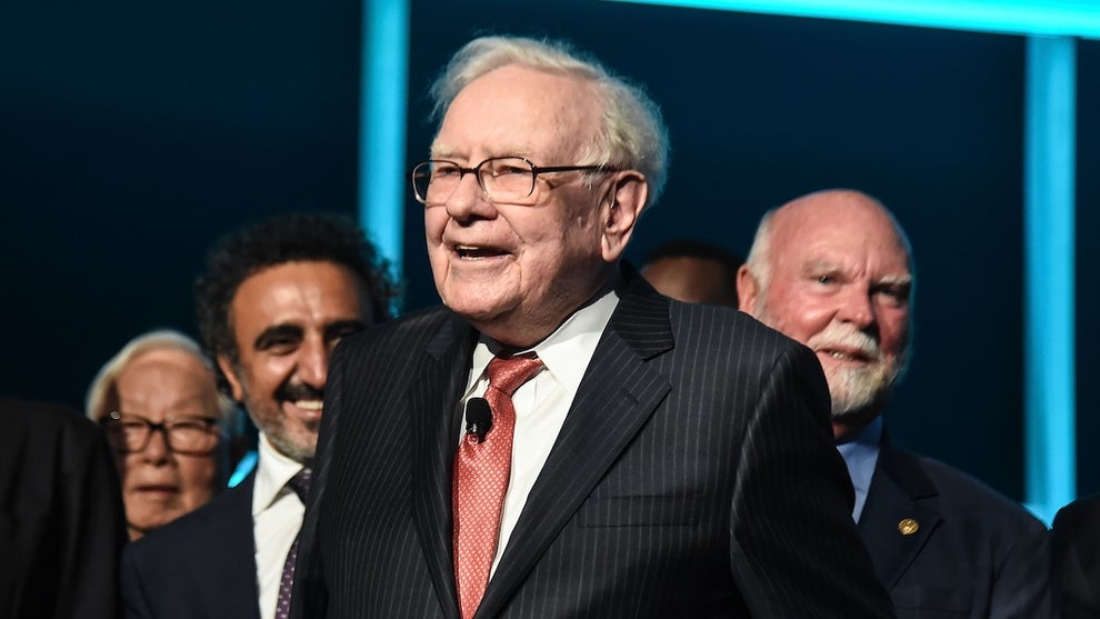 An older man with light-toned skin surrounded by other people also wearing suits. The man in the center is Warren Buffett.