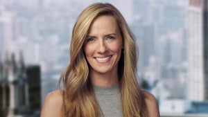 A blonde woman with light-toned skin smiles and poses in front of a cityscape behind her in soft focus.