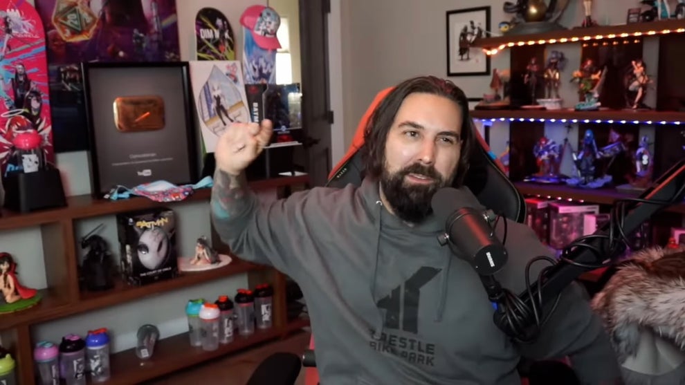 A light-skinned man with a beard and brown hair in front of walls with shelves filled with memorabilia. He makes an animated motion as he speaks into a microphone.