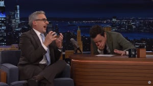 A man on the left wearing a suit gestures as a man on the right bends over laughing, sitting at a desk.
