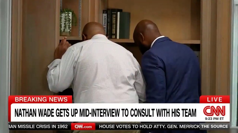 Two men speak as they face a bookcase, faces turned away from the camera. A chyron reads "NATHAN WADE GETS UP MID-INTERVIEW TO CONSULT WITH HIS TEAM." The two men are Black.