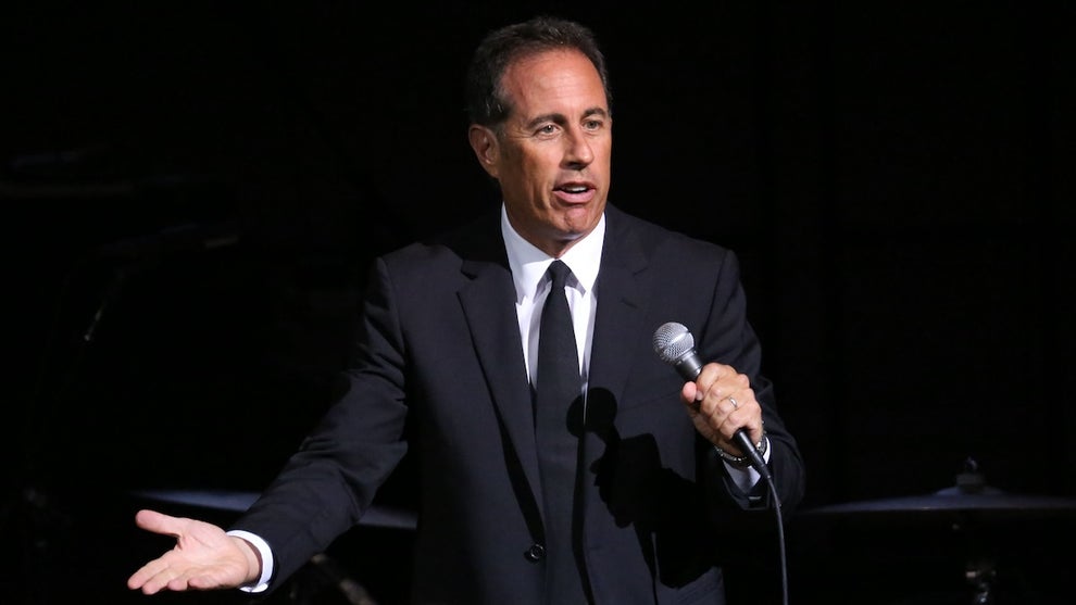 Jerry Seinfeld, a man with light-toned skin, looks incredulous as he holds a microphone and wears a suit, gesturing onstage.