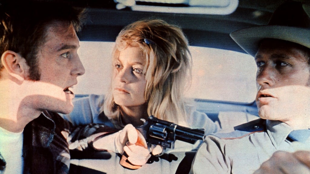 The man on the left holds a gun pointed toward the man on the right in a car, who looks afraid as he holds the wheel. A woman with blond hair sits in the back seat, looking at the man on the left.