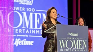 A woman in a long leather jacket speaks at a podium. She has light-toned skin and text on the podium reads The Hollywood Reporter, Power 100 Women in Entertainment.