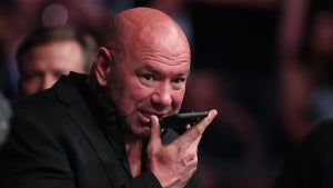 A bald man with light-toned skin speaks into a phone he holds in front of his face, with a large crowd in the background. The man is Dana White.