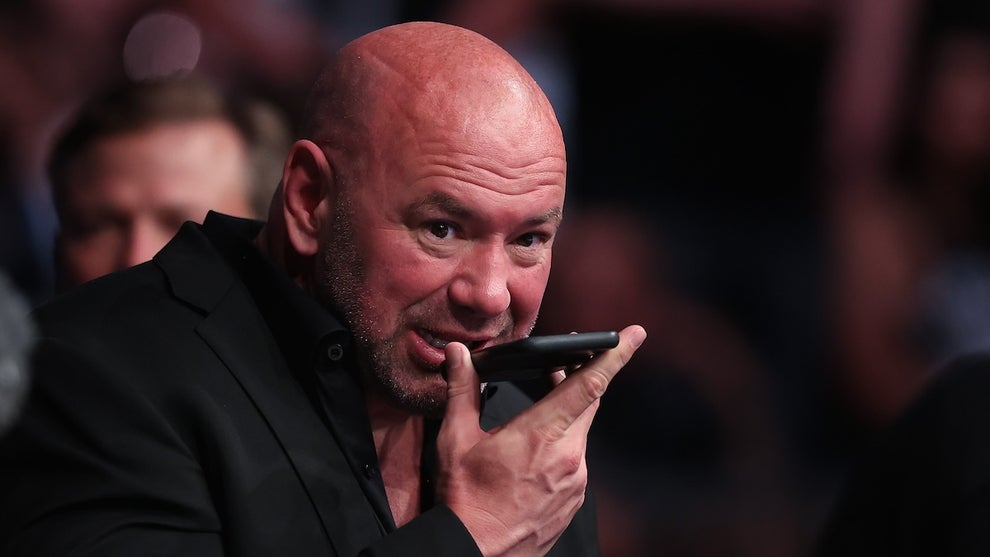 A bald man with light-toned skin speaks into a phone he holds in front of his face, with a large crowd in the background. The man is Dana White.