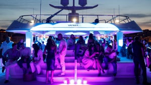 A purple neon-lit party aboard a yacht with a number of people mingling on its deck.