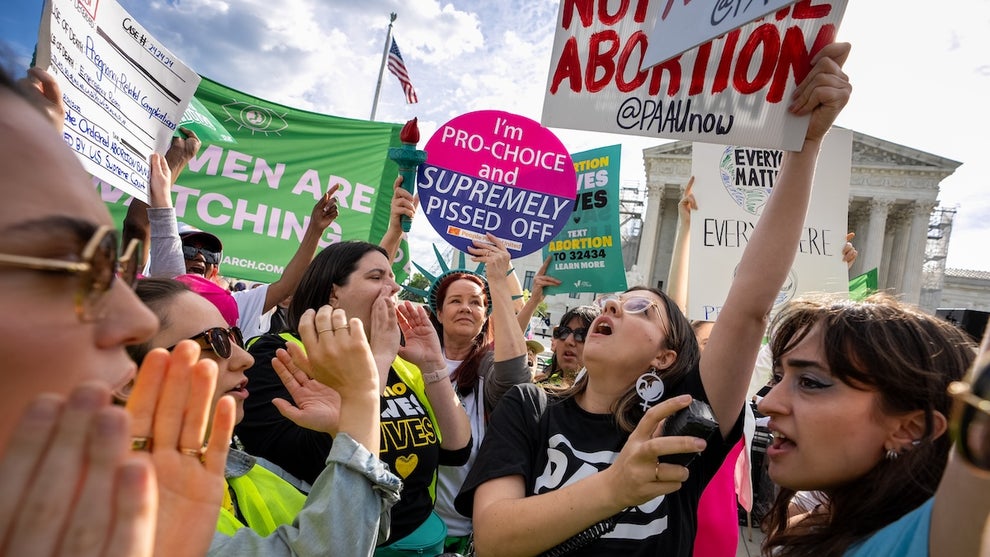 A crowd of protesters, mostly women, gather and yell at each other outside of the Supreme Court building. Many of them hold up signs with pro-choice slogans on them, while others cup their hands around their mouths to amplify their voices.