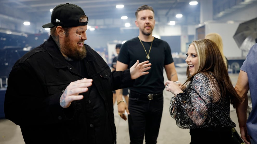 A large, smiling man with tattoos looks at a smaller, excited woman. They both have light-toned skin. Another man stands in the background.