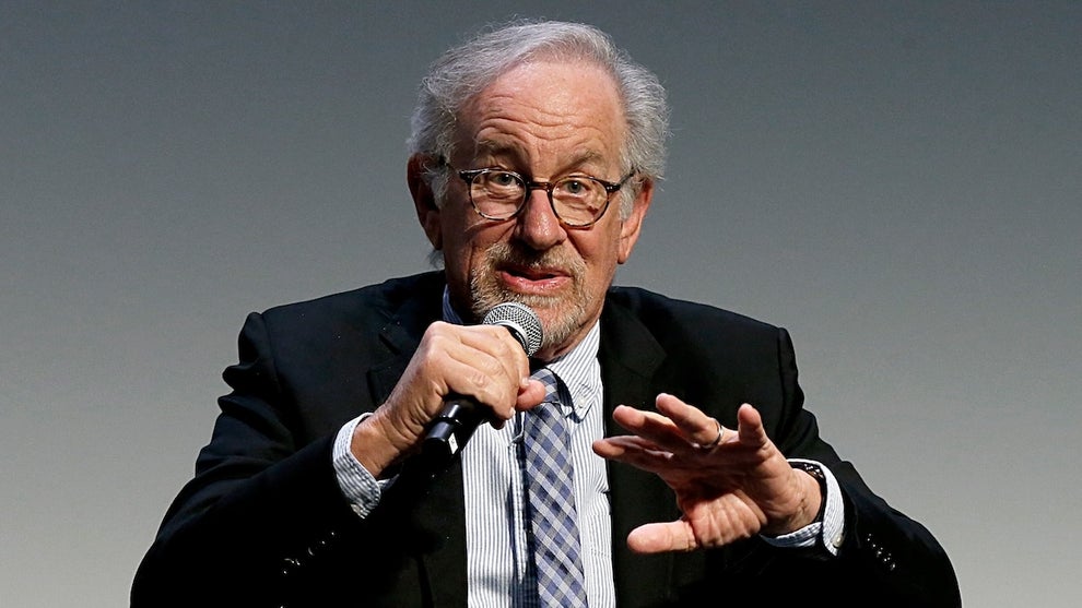 An older man with gray hair and a beard speaks into a microphone, wearing a sports coat, in front of a soft gray background.