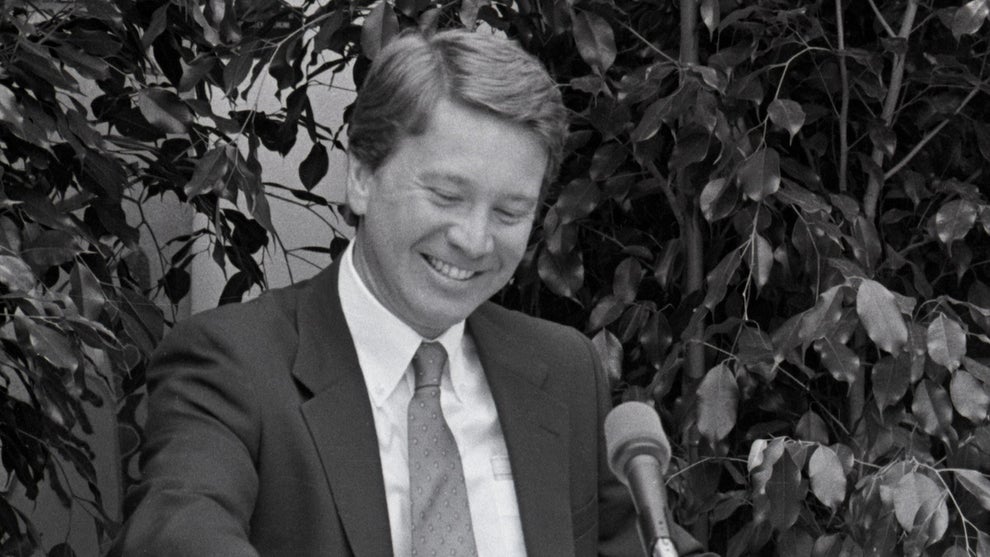 A man with light brown hair wears a suit as he sits in front of foliage, in front of a microphone at a press conference. The man with light-toned skin is Jamie Kellner.