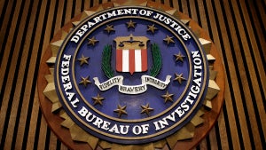 The Federal Bureau of Investigation's logo, which includes red and white stripes at the center, surrounded by laurels and stars, as well as the name of the organization and its parent department: Department of Justice, Federal Bureau of Investigation. A sash across the middle reads Fidelity, Bravery, Integrity. It is on a brown-paneled wall.