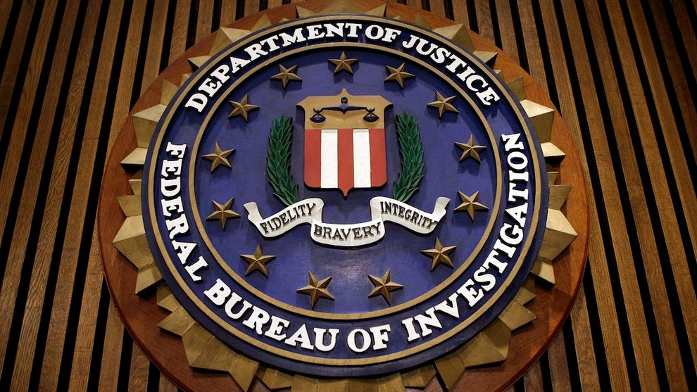 The Federal Bureau of Investigation's logo, which includes red and white stripes at the center, surrounded by laurels and stars, as well as the name of the organization and its parent department: Department of Justice, Federal Bureau of Investigation. A sash across the middle reads Fidelity, Bravery, Integrity. It is on a brown-paneled wall.