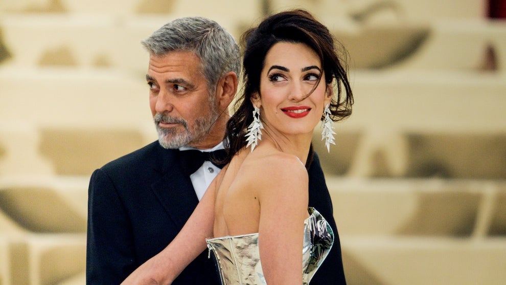 An older man stands with a woman, touching her lower back. He wears a tuxedo while she wears a silver dress. He is tan but with light-toned skin, while she has medium-toned skin.