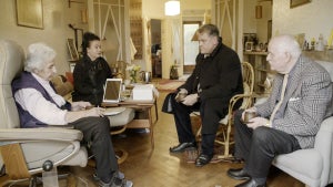 A group of older light-skinned men and women sit across from one another.
