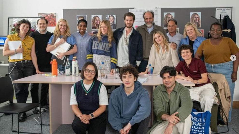 A group shot of a number of actors around a table in a relatively bare room.