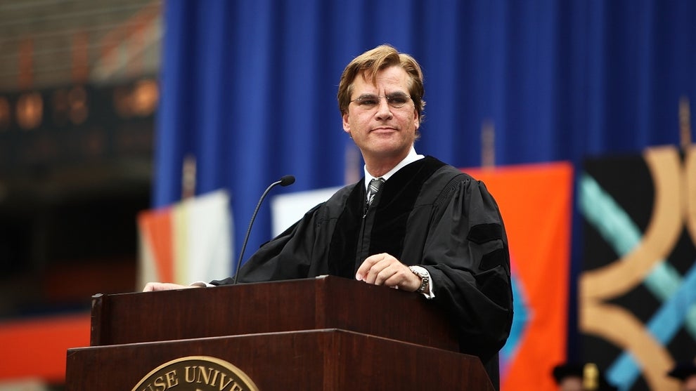 A man with light-toned skin wearing a black academic robe stands at a lectern on stage with a blue backdrop behind him.