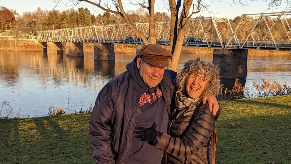 An older man and woman stand outside in front of a scenic bridge in the background.