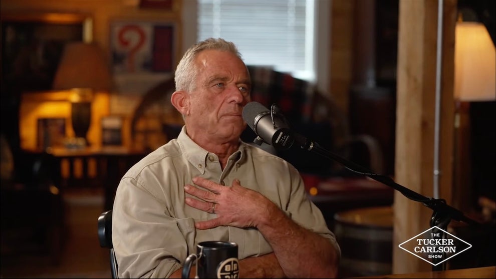 An older man puts his hand to his chest, gesturing as he sits in a cabin in front of a microphone.
