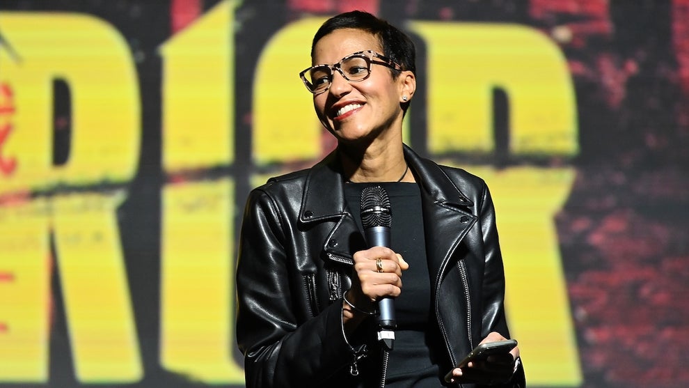 A woman with medium-toned skin stands on stage, smiling and holding a microphone in front of a backdrop reading "Warrior."