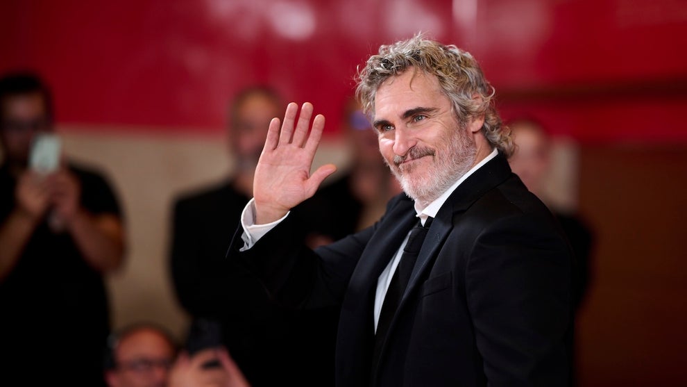 A middle-aged actor with graying hair waves, wearing a tuxedo on a red carpet.