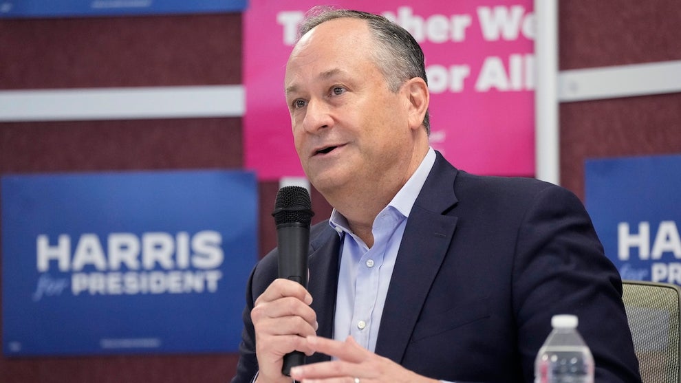 A man with light-toned skin in front of a group of pink and blue signs. The blue signs read "Harris for president." He wears a sport coat and holds a microphone. A bottle of water is on the table in front of him.