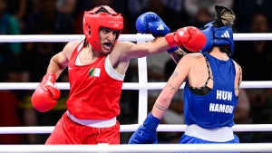 Boxer Imane Khelif wears Red, throwing a cross against her opponent in blue in the middle of a boxing ring, a crowd seated and watching in the background.