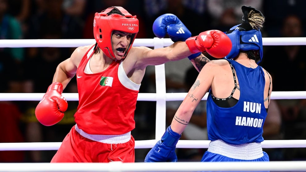 Boxer Imane Khelif wears Red, throwing a cross against her opponent in blue in the middle of a boxing ring, a crowd seated and watching in the background.