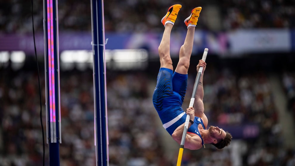 An upside-down pole vaulter, mid-vault, wearing a blue singlet with a crowd watching behind him.
