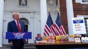 A man in a suit stands in front of a podium next to a table full of common groceries and American flags in front of a large building with pillars.
