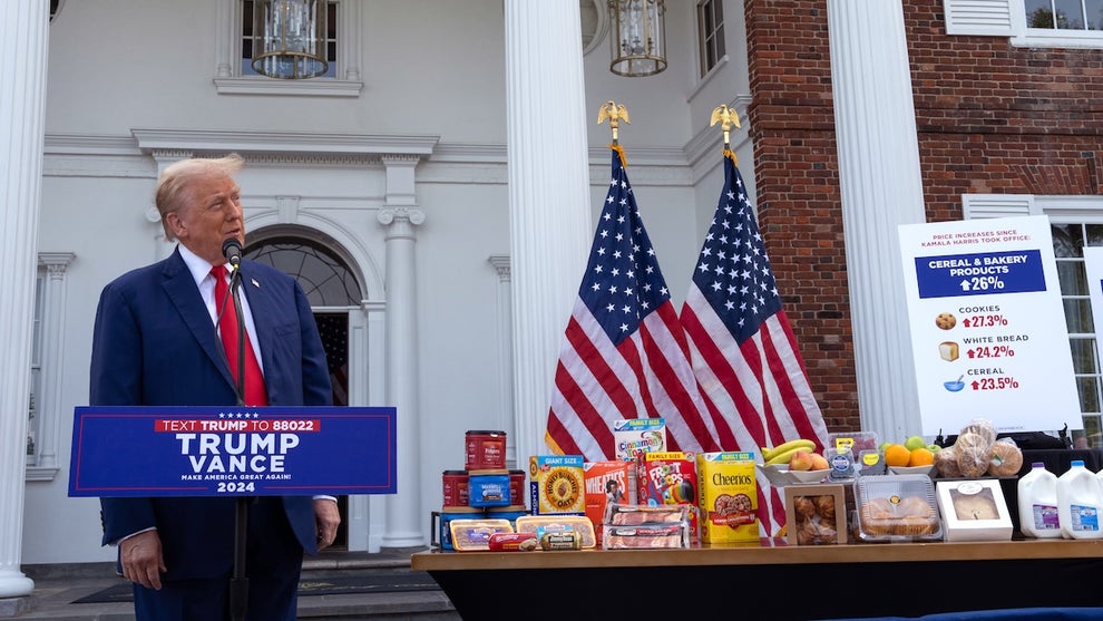 A man in a suit stands in front of a podium next to a table full of common groceries and American flags in front of a large building with pillars.