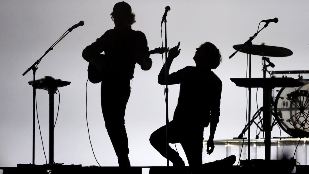 Two men perform on stage, one playing a guitar and the other kneeling with a microphone, shown in silhouette with a drum kit and mic stands also featured.