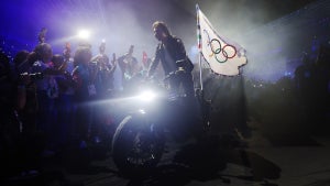 Tom Cruise, a man with light-toned skin, rights a motorcycle with the Olympics flag showing the Olympic rings on it on the back, surrounded by a crowd in a stadium.