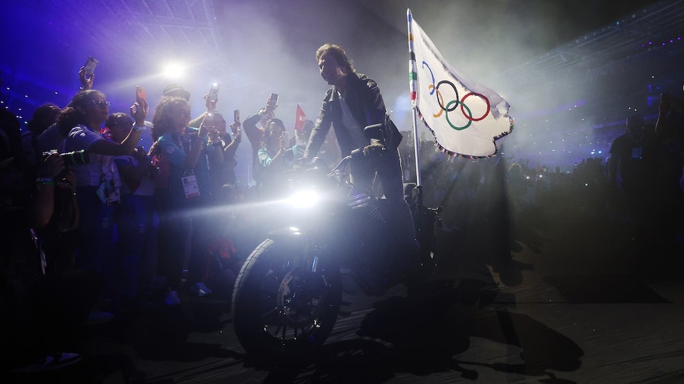 Tom Cruise, a man with light-toned skin, rights a motorcycle with the Olympics flag showing the Olympic rings on it on the back, surrounded by a crowd in a stadium.