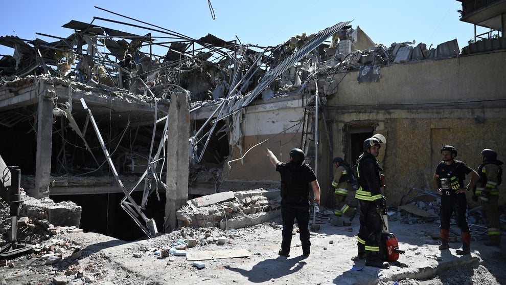 The wreckage of a mid-sized building that appears to be a couple stories tall, with several people standing and pointing as they work on it following the destruction.