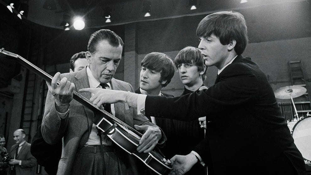 A group of five men, with an older man holding a guitar and wearing a suit while another illustrates how to use it.