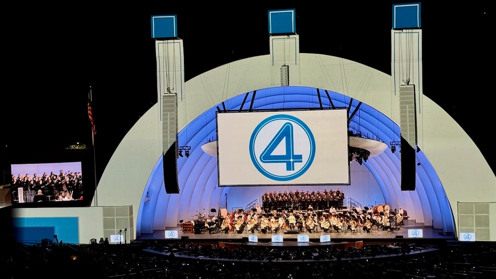 A large stage at the Hollywood Bowl, the arch illuminated in white with a projection screen with a Fantastic 4 logo, a blue 4 in a circle. On stage are an orchestra and a choir, playing at night in front of a packed crowd.
