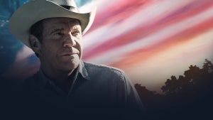 An image of a man in a cowboy hat in front of a red, white, and blue background that looks like the American flag in an impressionistic sense in the sky.