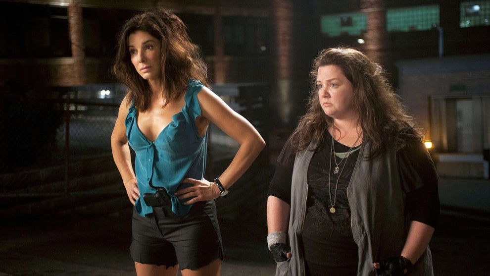 Sandra Bullock and Melissa McCarthy stand outside at night in a scene from The Heat (2013). Bullock wears a teal sleeveless blouse and black shorts, hands on hips with a serious expression, while McCarthy, dressed in layered dark clothes and fingerless gloves, stands beside her looking unimpressed. The setting is dimly lit with industrial buildings in the background, suggesting a tense or comedic stakeout moment.