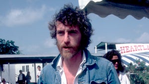 A man with a beard and curly hair, wearing a jean jacket. He has light-toned skin and is standing outside in a boardwalk-esque area.