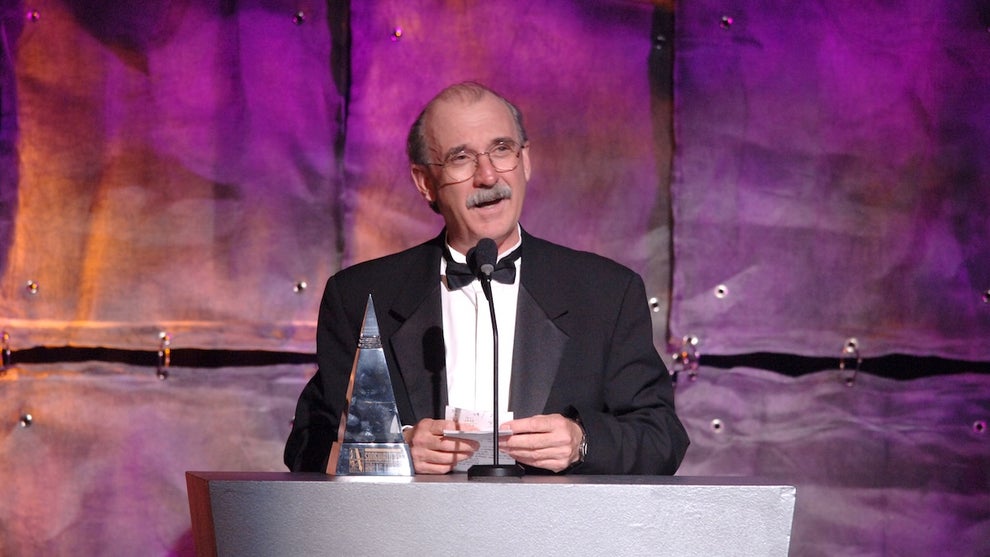 An older man wearing a tuxedo stands in front of a microphone and podium accepting an award on stage, in front of a textured background.