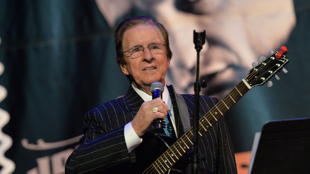 An older main in a suit holds a microphone and has a guitar slung around his body, standing in front of a mic stand. In the background is a large photo on a backdrop of his brother, Johnny Cash. Both men have light-toned skin.