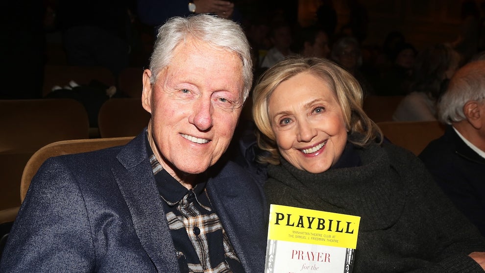 An older man and woman sit in a theater. The man holds up a playbill. They both have light-toned skin.
