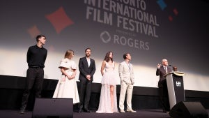 A group of people stand on a stage; in the background, a movie screen has a projection that reads Toronto International Film Festival. There are six people on stage, including two women and four men. They all have light-toned skin.