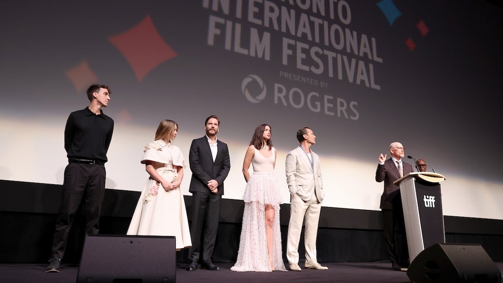 A group of people stand on a stage; in the background, a movie screen has a projection that reads Toronto International Film Festival. There are six people on stage, including two women and four men. They all have light-toned skin.
