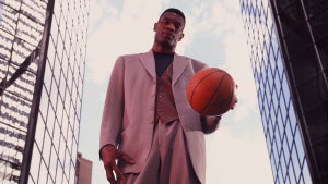 A man with dark-toned skin stands in a suit, holding a basketball, in between two skyscrapers.