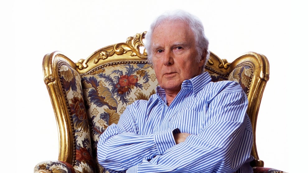 An older man sits, arms crossed, in a fanciful gold-trimmed chair.